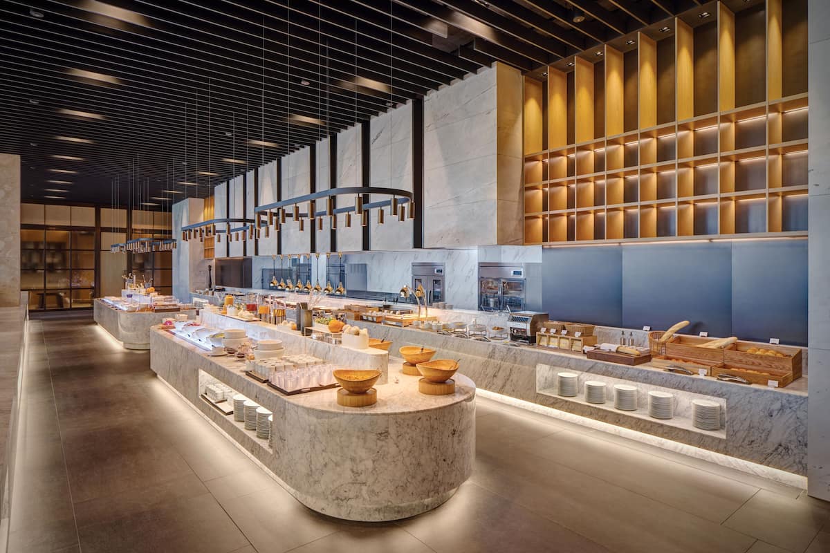 A modern, well-lit hotel lobby with a marble reception desk, a variety of food and beverage stations, and a large seating area.