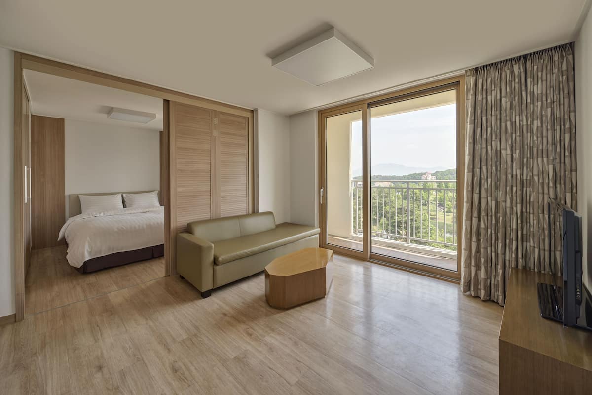 A modern hotel room with a bed, a sofa, a small table, and a large sliding glass door leading to a balcony with a view of the city.