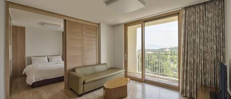 A modern hotel room with a bed, a sofa, a small table, and a large sliding glass door leading to a balcony with a view of the city.