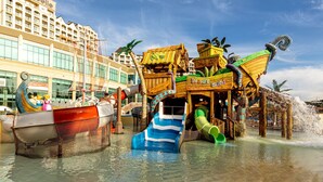 Un parc aquatique avec une attraction proposant un parcours en bateau en bois, un toboggan bleu et vert, et une fontaine d’eau.