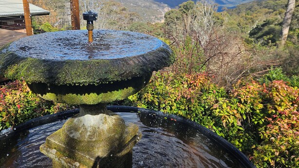 A stone fountain with water spouting from the top, surrounded by lush greenery and a view of distant hills. - STUNNING VIEWS, 6 bedrooms, 12 people, fireplace, early check-in, late check-out (Mount Victoria)