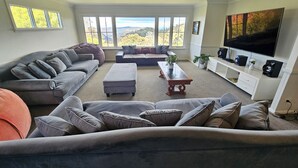 A spacious living room with a grey sectional sofa, a smaller sofa, a coffee table, and a TV on a white cabinet. Large windows provide a view of the outdoors. - STUNNING VIEWS, 6 bedrooms, 12 people, fireplace, early check-in, late check-out (Mount Victoria)