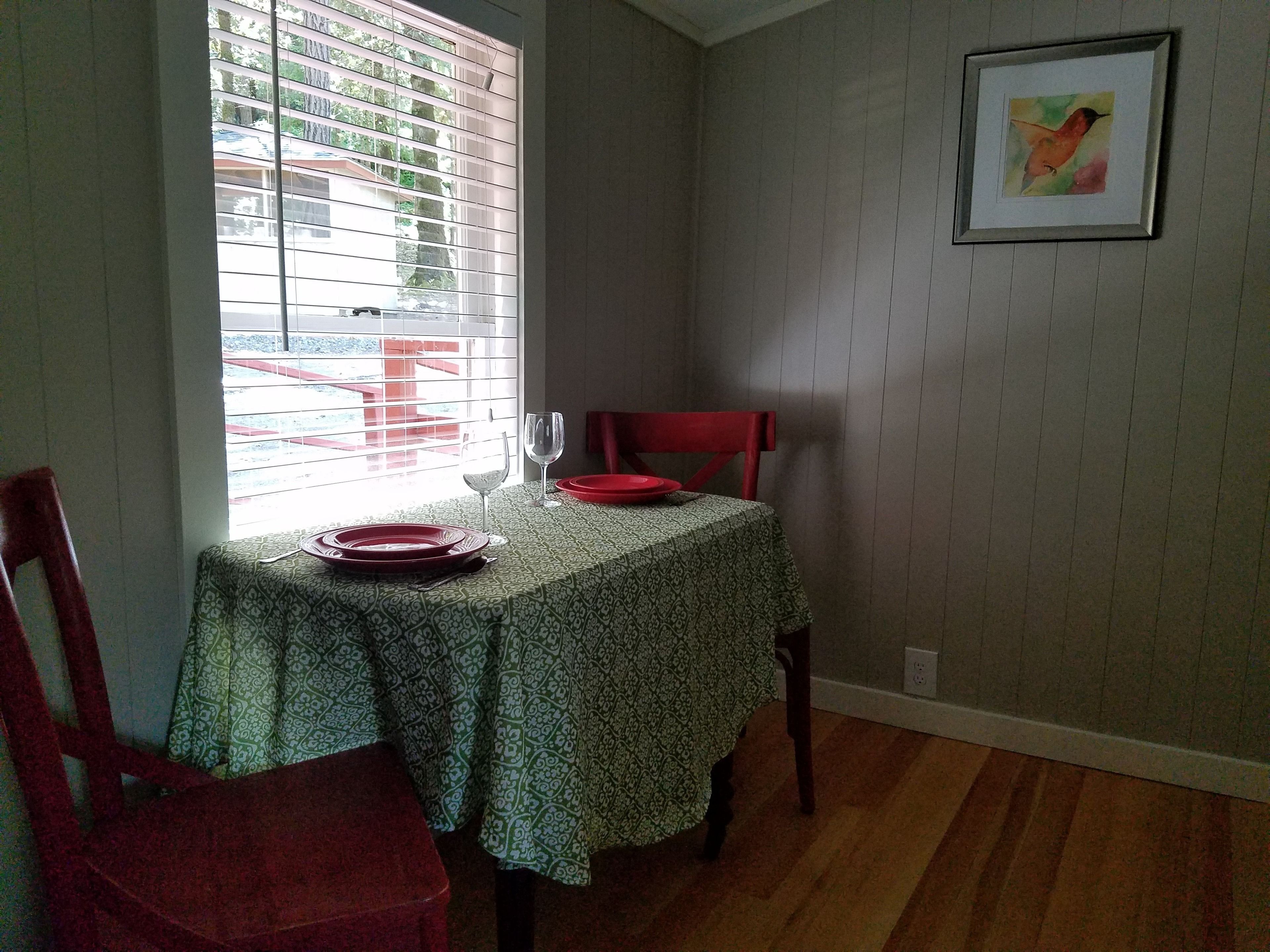 A dining area with a table set for two, a window with blinds, a red chair, and a framed picture on the wall.