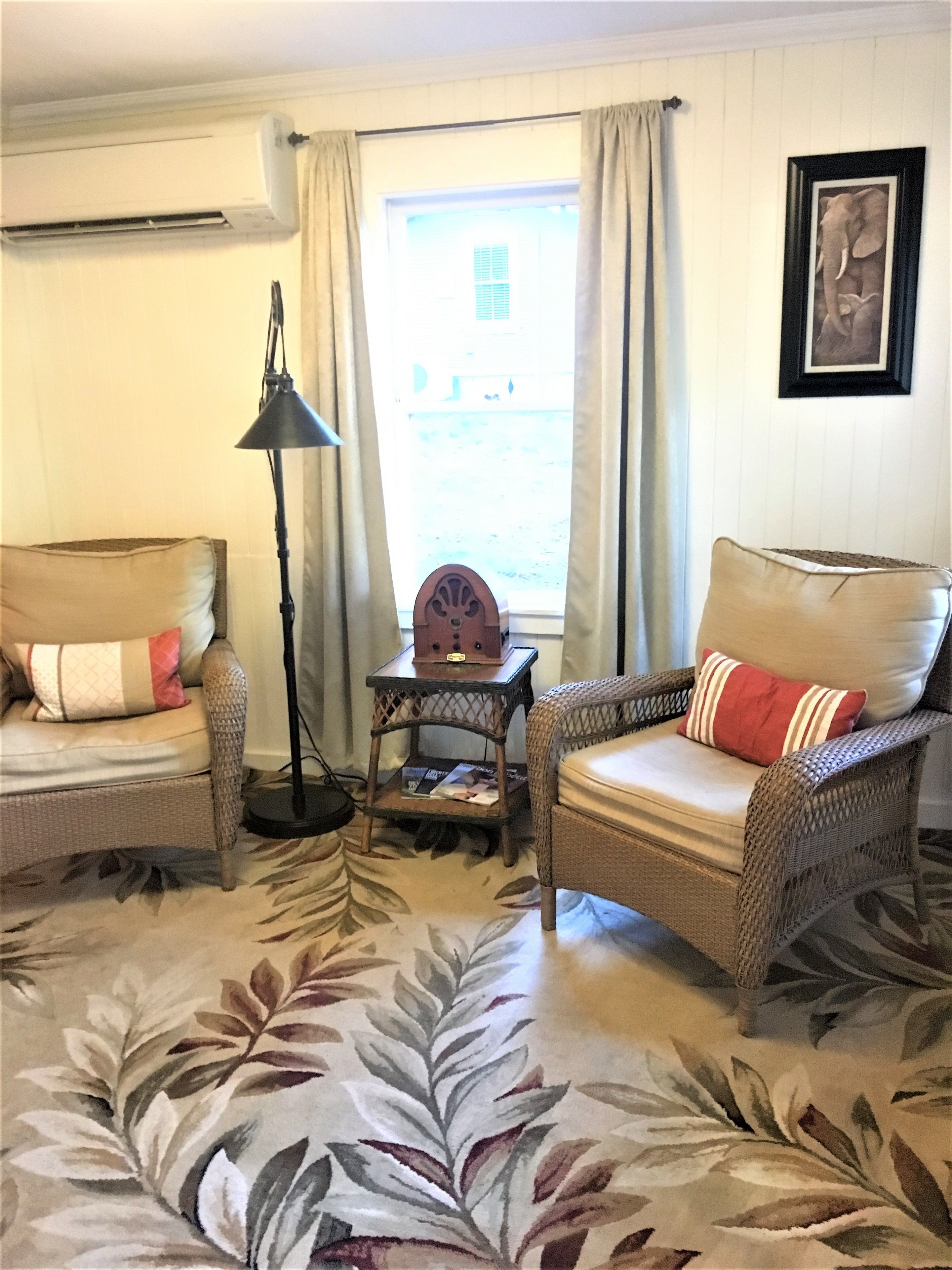 A living room with two wicker chairs, a floor lamp, a small table with magazines, and a window with curtains.