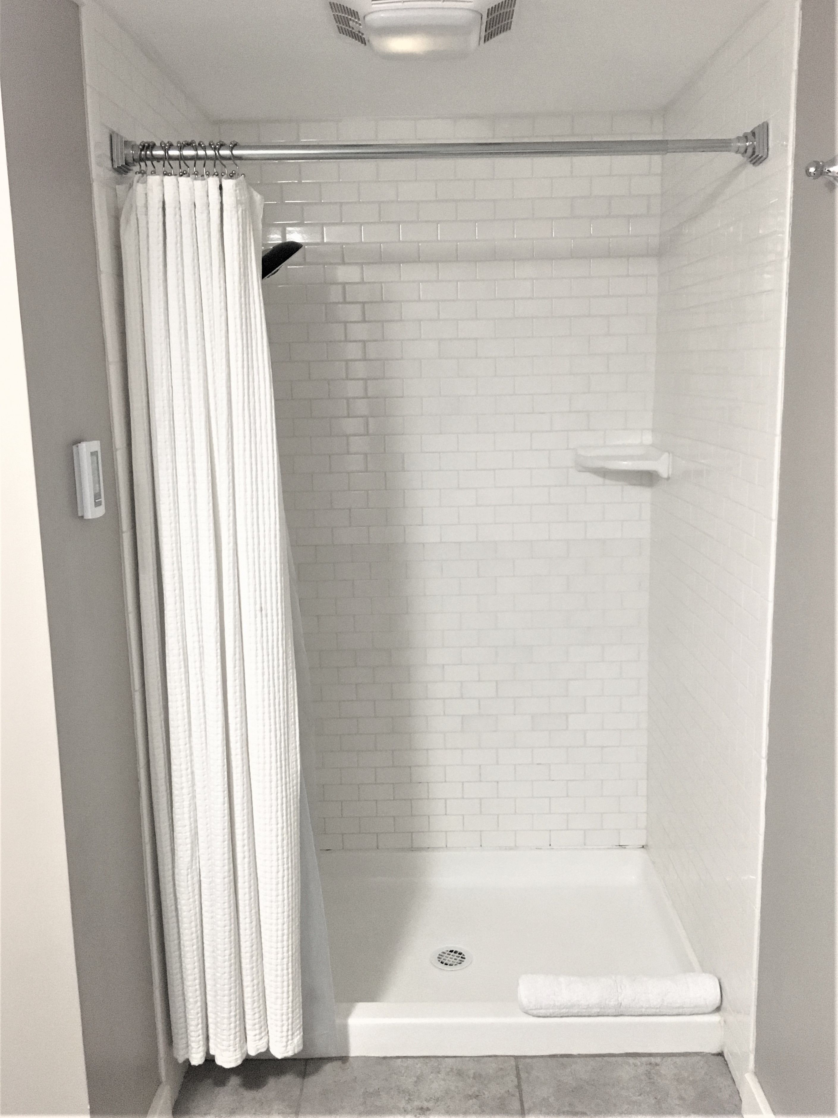 A white-tiled shower enclosure with a shower curtain, a handheld showerhead, and a small shelf.