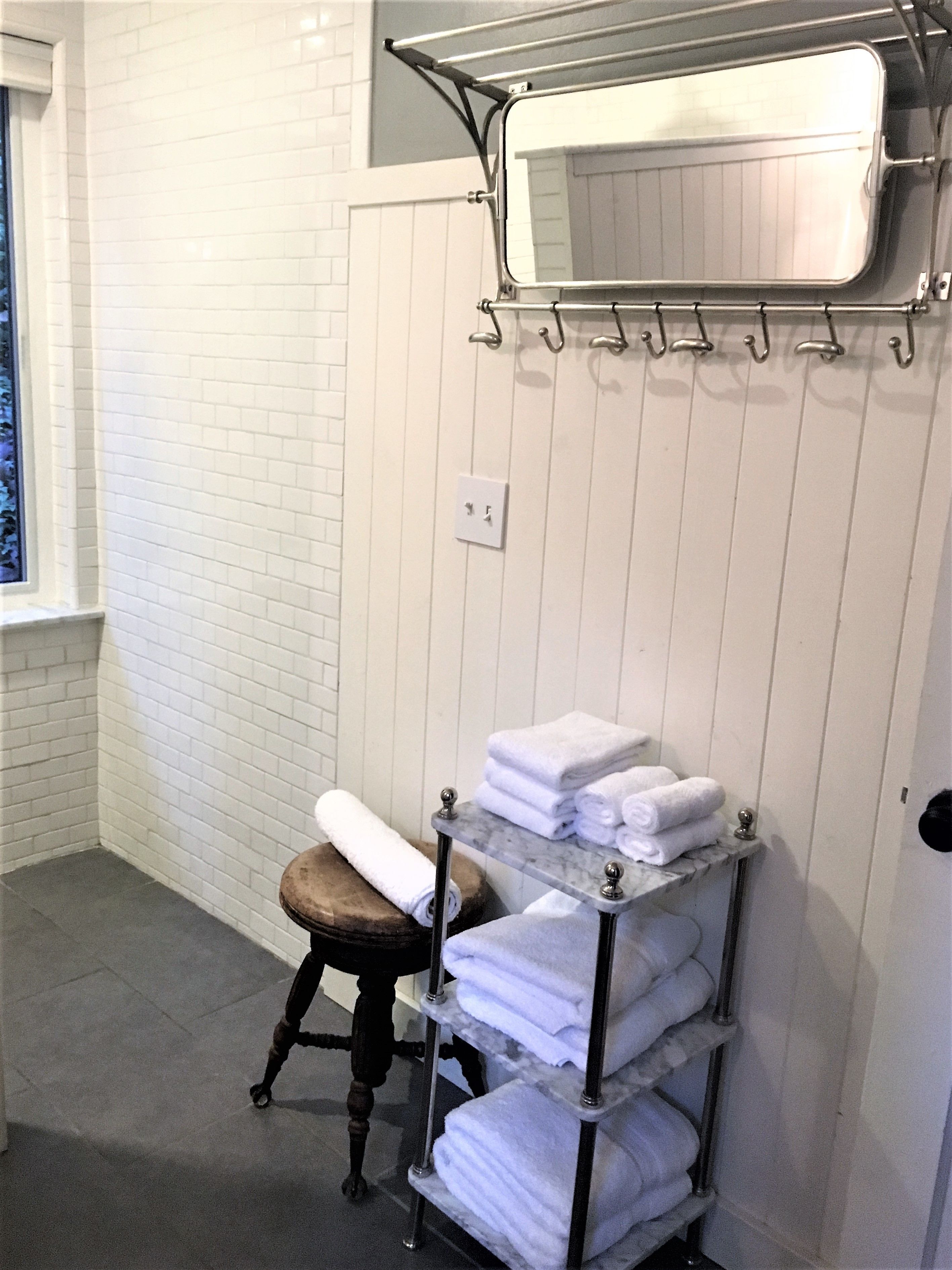 A bathroom with a white tiled wall, a wooden stool, a metal towel rack with folded towels, and a mirror above the rack.