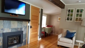 A living room with a fireplace, a television, a dining area with a table and chairs, and a white armchair. - China Creek Cottages (Willow Creek)