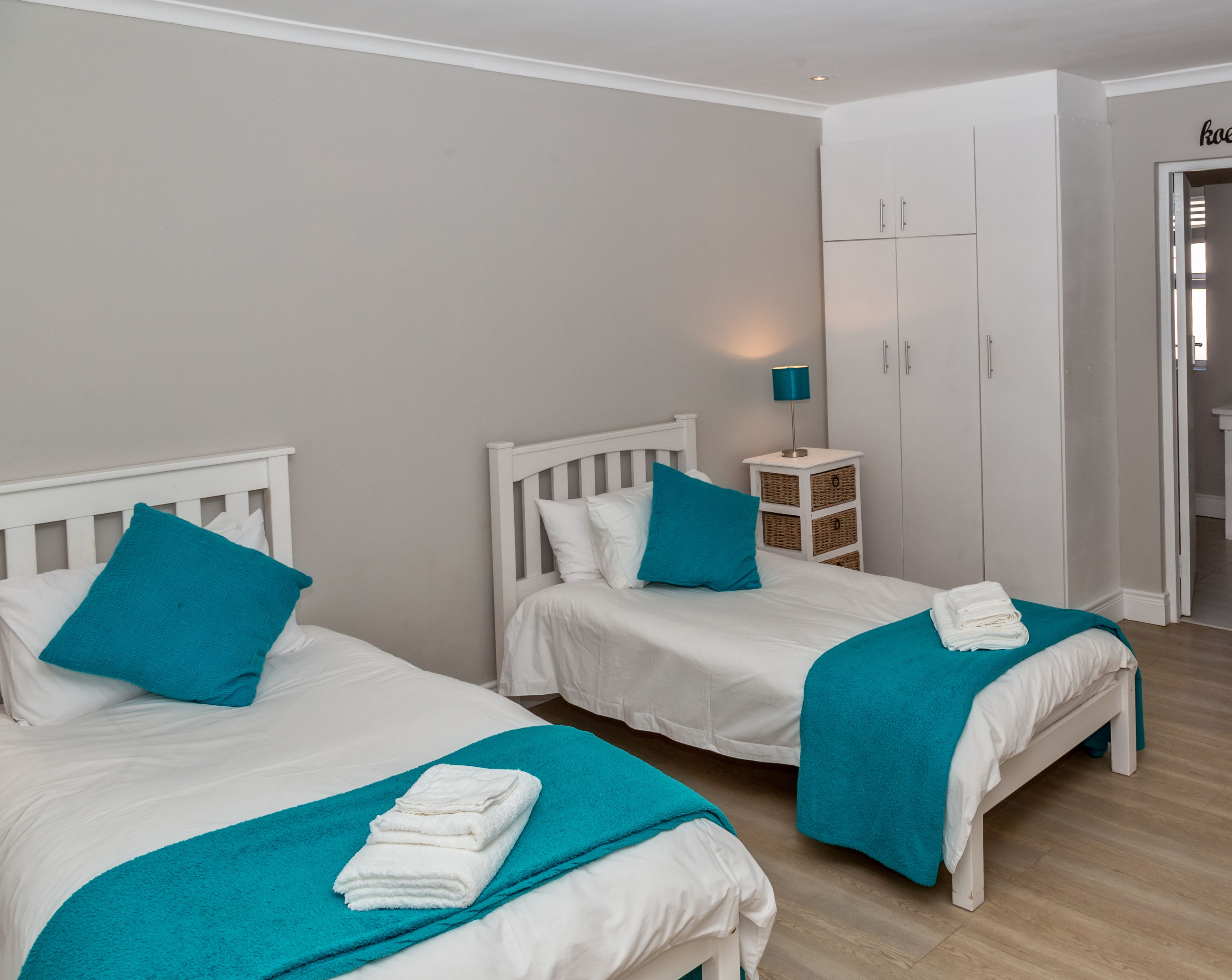 A bedroom with two beds, each with white bedding and teal accents, a small bedside table with a lamp, and a wardrobe.