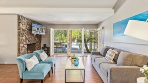 A spacious living room with a stone wall, two sofas, a coffee table, and a view of an outdoor patio with a pool and palm trees. - Private Pool Resort Home - Walk to Music Festivals Polo Grounds (Indio)