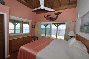 A bedroom with a large bed, a dresser, a ceiling fan, and a view of the ocean through a sliding door.