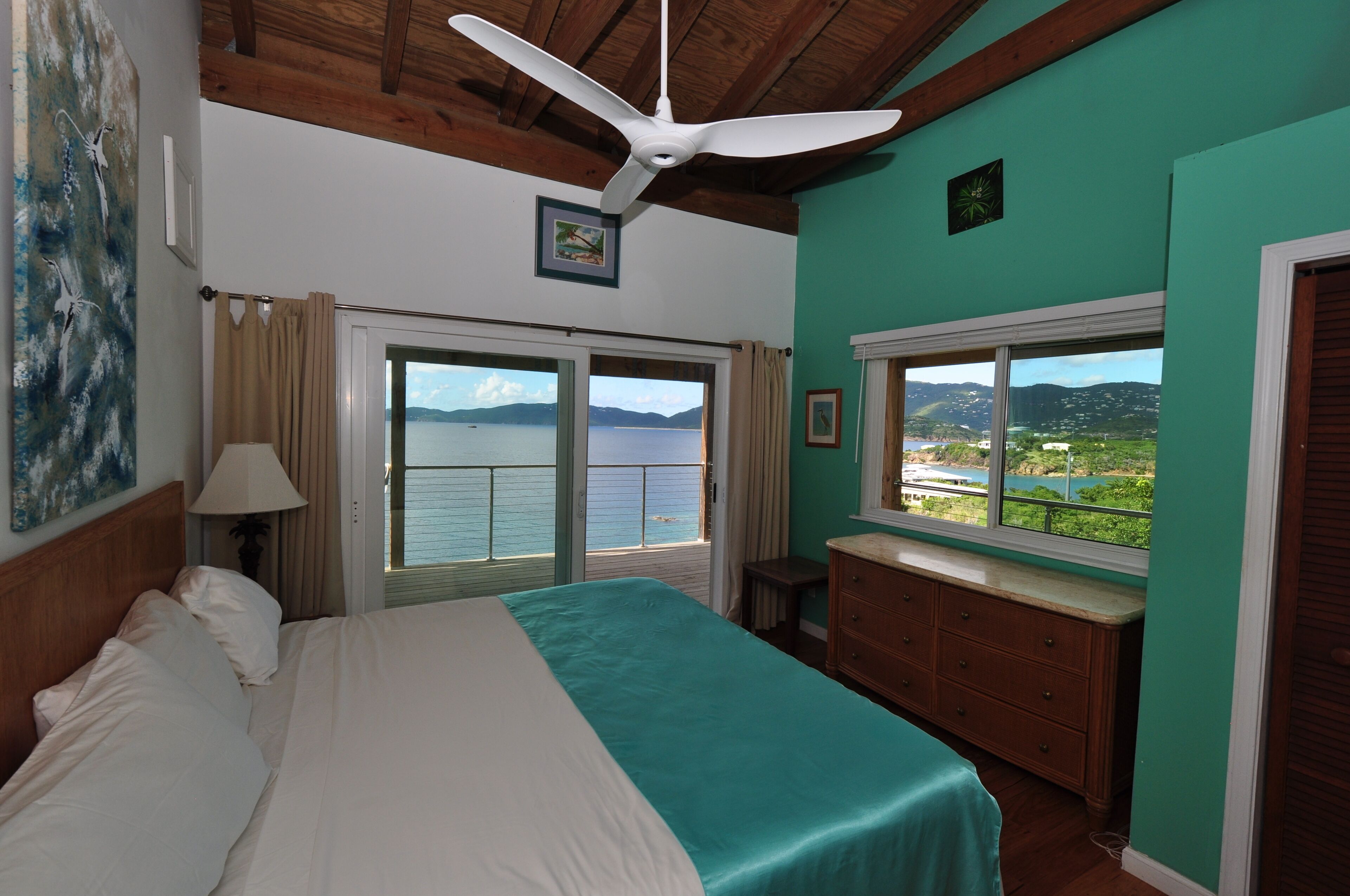 A bedroom with a bed, a ceiling fan, a window with a view of the sea, and a dresser.