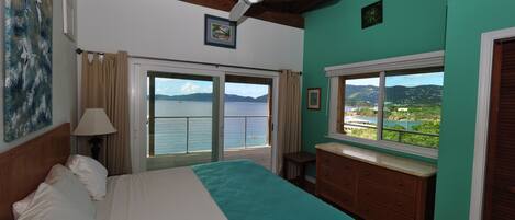 A bedroom with a bed, a ceiling fan, a window with a view of the sea, and a dresser.