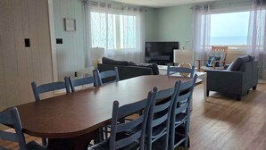 A dining area with a wooden table and blue chairs, a living room with a sofa and TV, and a view of the sea through large windows. - 3 bedroom 2 bathroom, pet friendly, ocean front duplex (Emerald Isle)