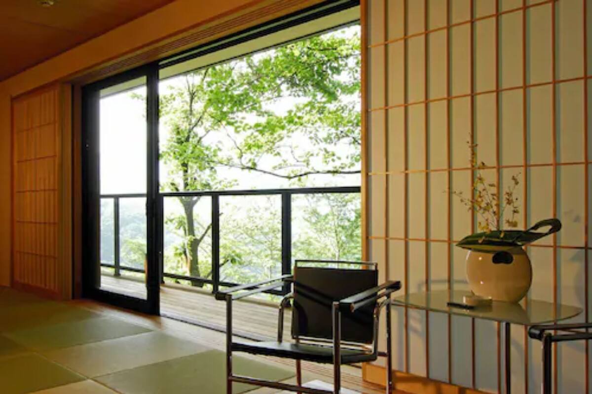 Una habitación con suelo de tatami, un fusuma que da a un balcón con vistas a árboles, una silla negra y un jarrón sobre una mesa de cristal.