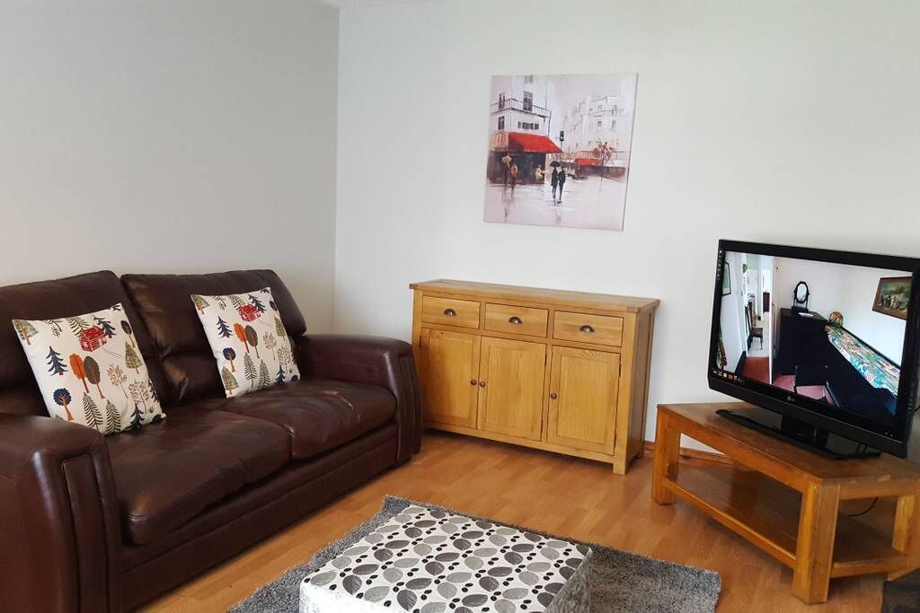 Een woonkamer met een bruine leren bank, een houten kast, een televisie op een houten dressoir en een kamerbreed tapijt met een patroon.