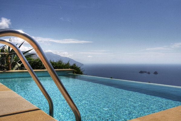 An infinity pool with clear blue water, a metal pool ladder, and a view of the ocean and distant land.