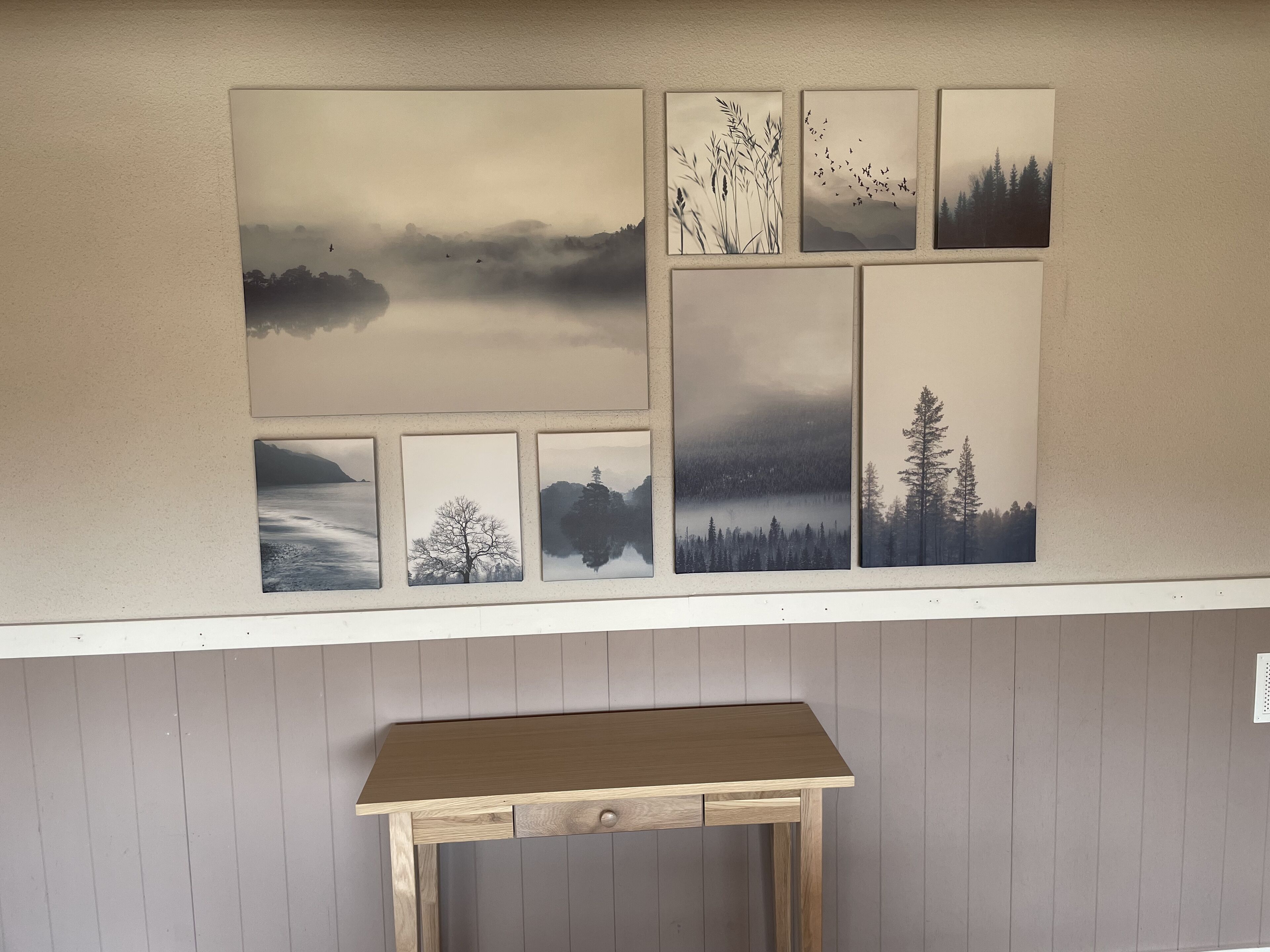 Eine Wand mit einer Reihe gerahmter Landschaftsfotografien und einem kleinen Holztisch darunter.