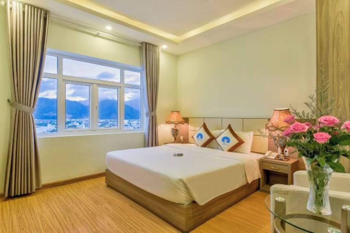 Een hotelkamer met een groot raam, een bed, bedlampen, een nachtkastje en een vaas met bloemen.