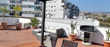 A rooftop terrace with outdoor furniture, potted plants, and a grill.