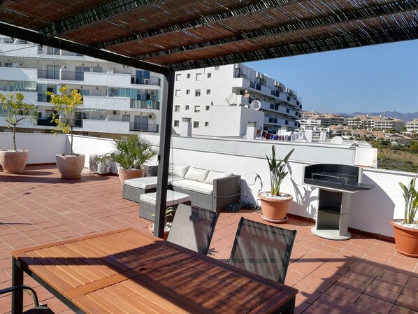 Una terraza en la azotea con mobiliario exterior, plantas en macetas y una parrilla.
