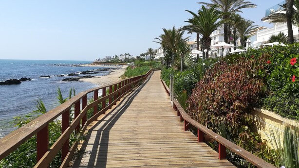 Eine hölzerne Promenade führt zu einem Strand mit Palmen und einem weißen Gebäude im Hintergrund.