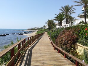 Un paseo marítimo de madera que conduce a una playa con palmeras y un edificio blanco al fondo.