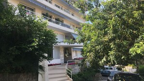 Un edificio de varios pisos con balcones, una escalera que conduce a la entrada y abundante vegetación alrededor.
