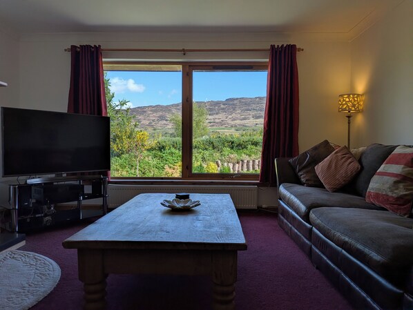 A living room with a television, a coffee table, a sofa, and a view of a mountainous landscape.