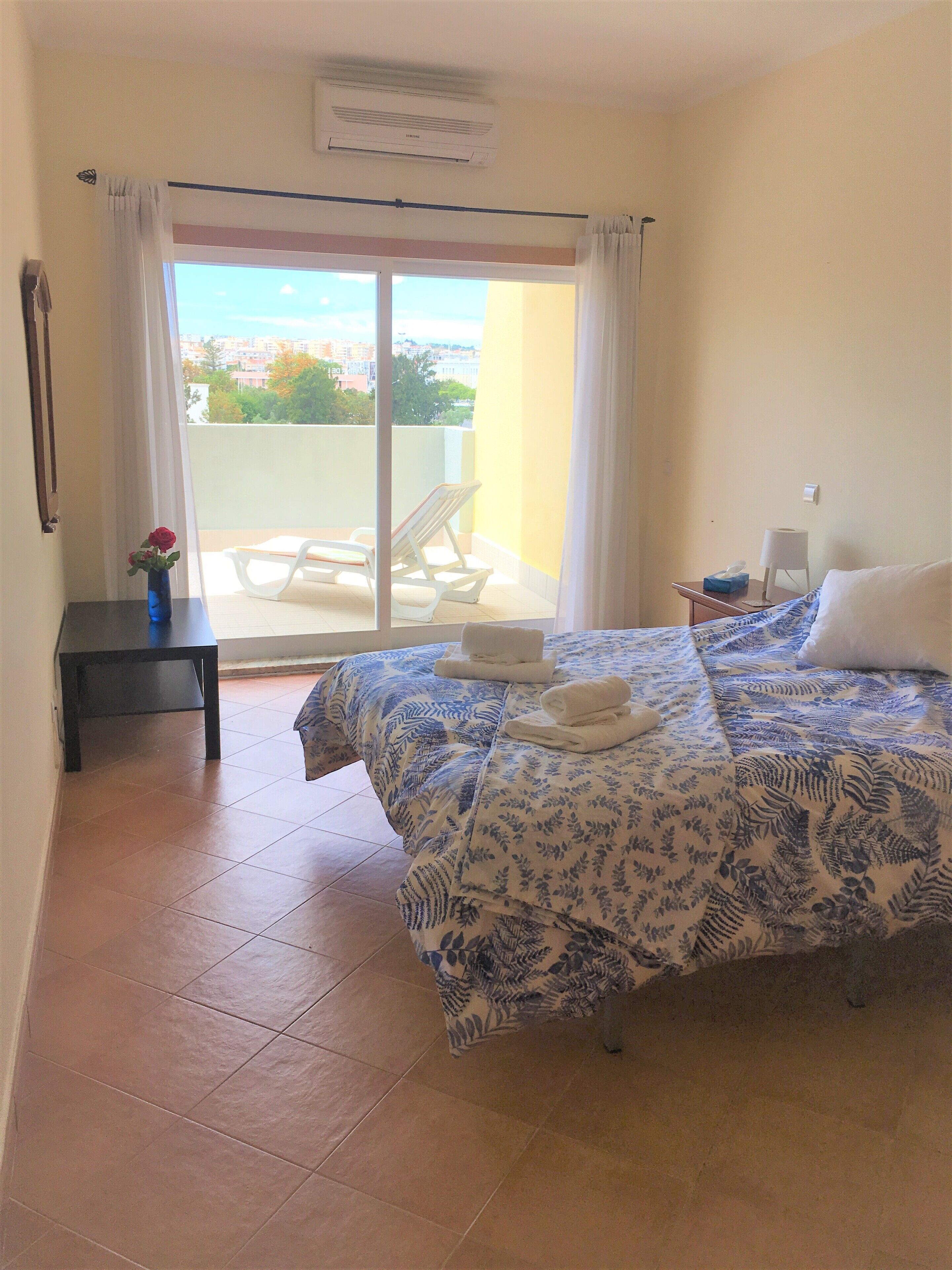 Quarto de hotel com uma cama, uma mesinha com um vaso de flores, uma mesaninha de cabeceira, um abajur e vista da cidade através de uma porta de varanda.