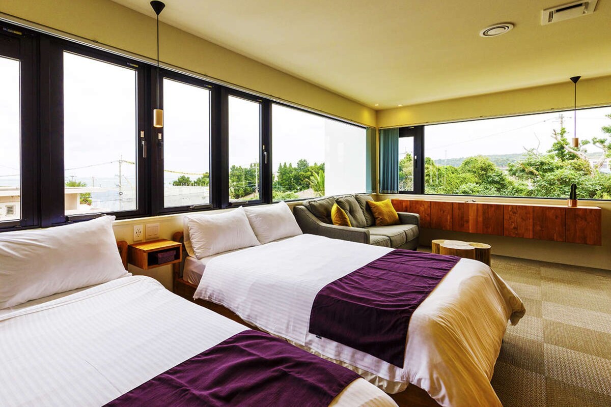 Ein Hotelzimmer mit zwei Betten, einem Sofa, einer hölzernen Bank und großen Fenstern mit Blick auf Grünflächen und Gebäude.