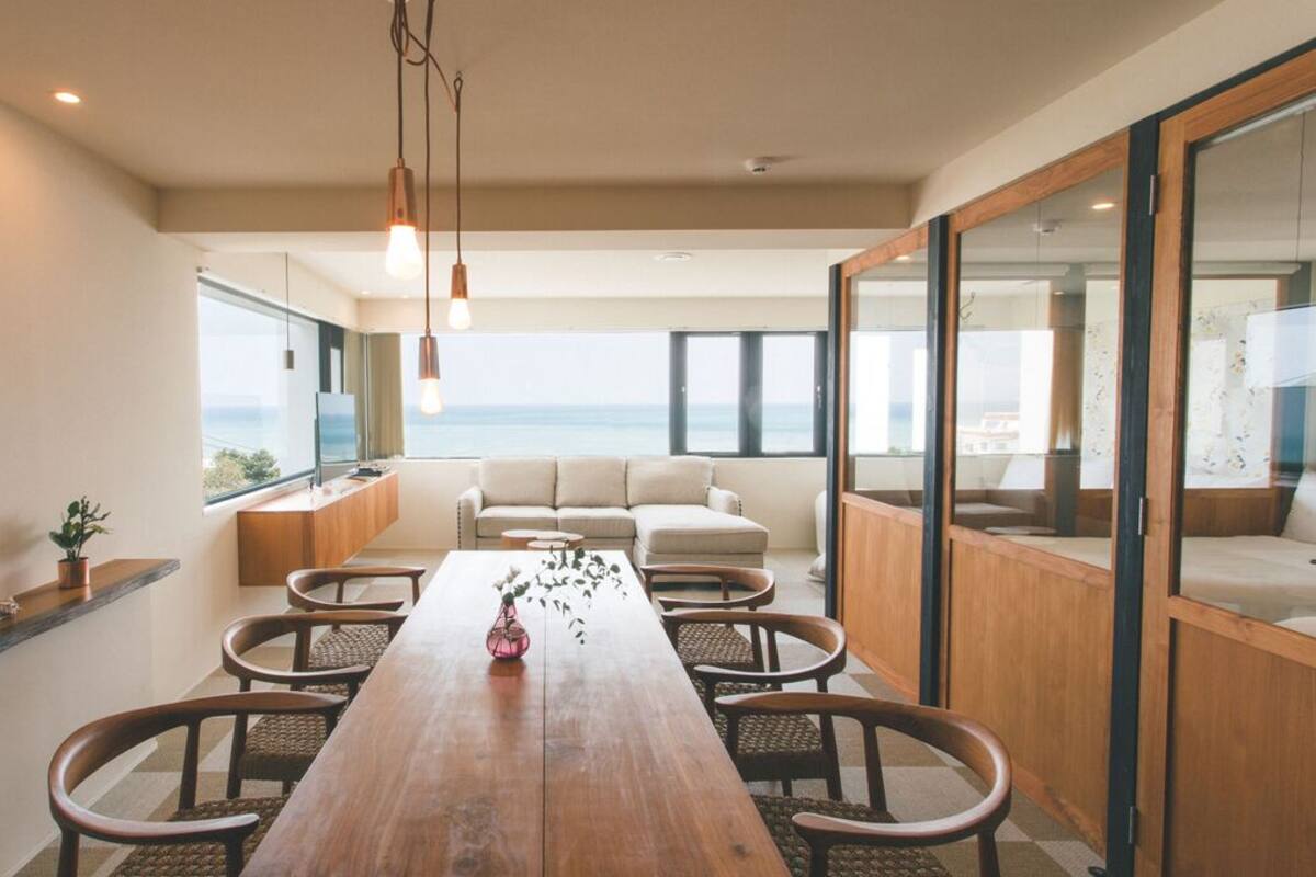 A modern dining area with a wooden table, chairs, and pendant lights. There is a view of the sea through large windows.