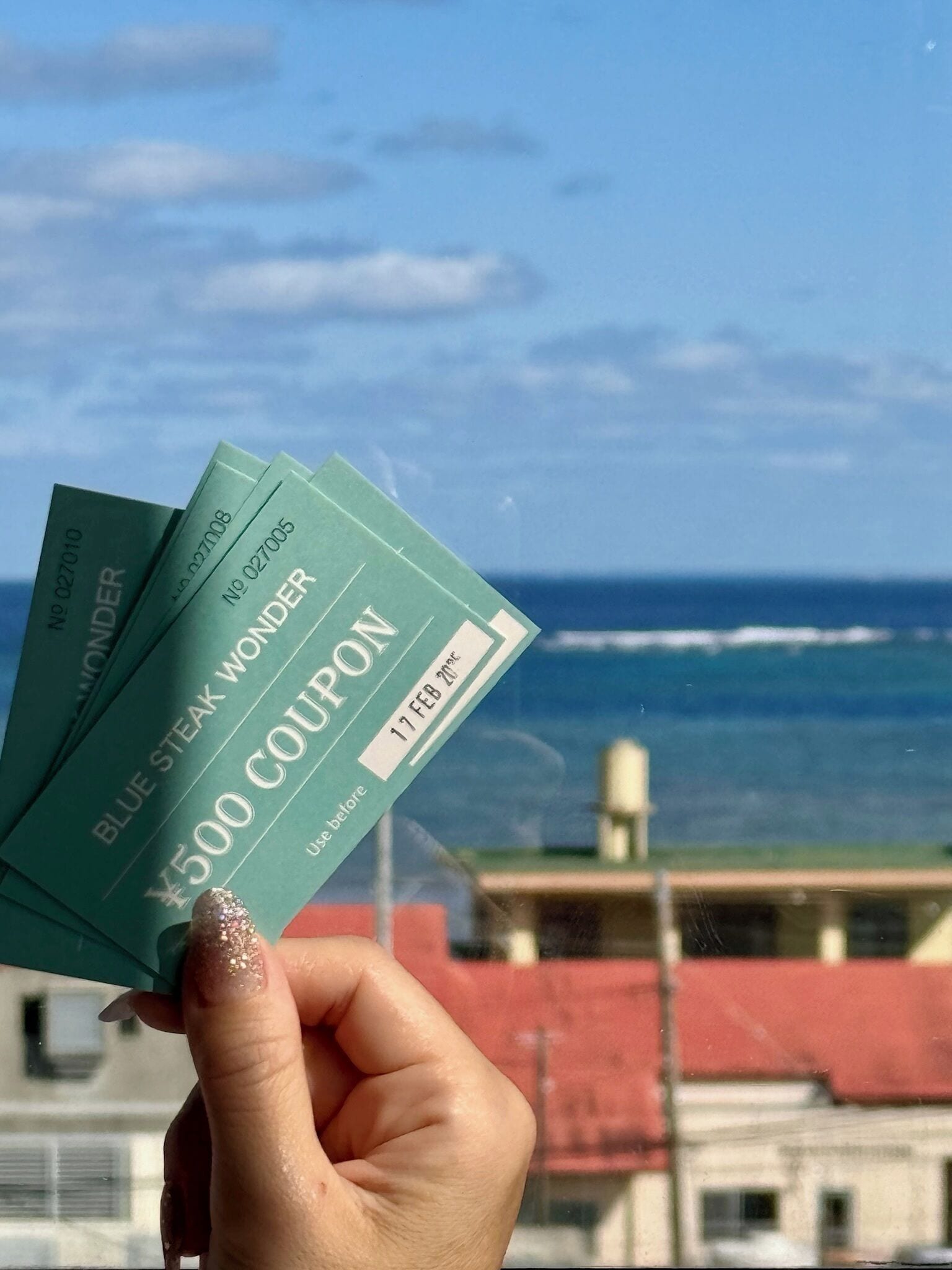 A hand holding three blue coupons with the text "Blue Steak Wonder" and "¥500 Coupon" against a backdrop of a building and ocean view.