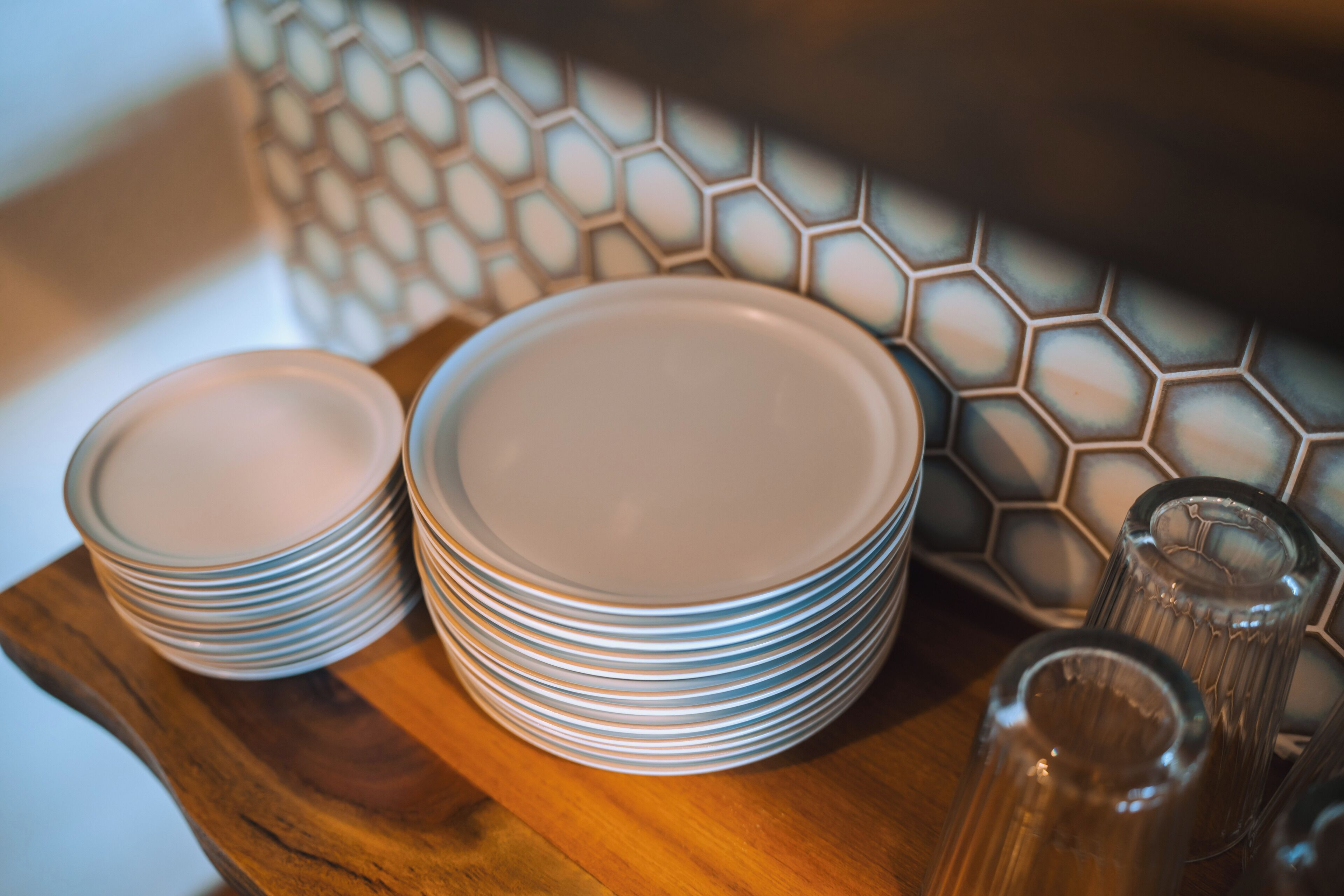 A stack of white plates on a wooden tray, a clear glass bottle, and a hexagonal patterned tile backsplash.