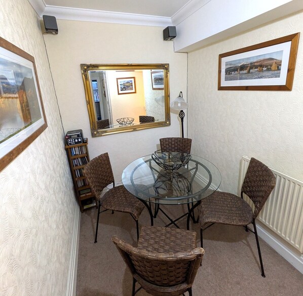 A dining area with a glass table surrounded by wicker chairs, a large mirror, and framed pictures on the walls. - A 2 bedroomed apartment in the middle of Keswick with fantastic mountain views (keswick)