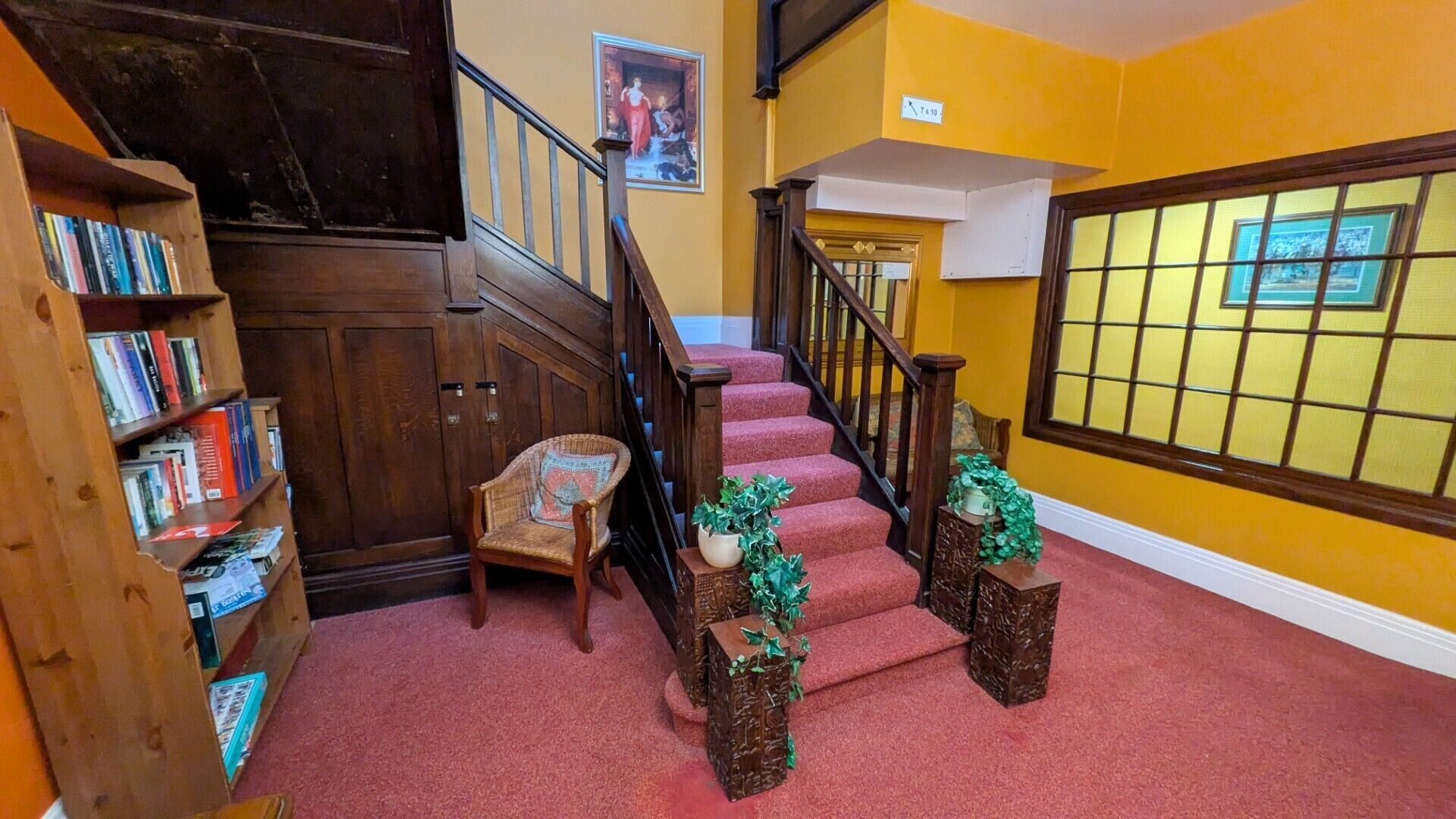 A staircase with red carpet, wooden railings, a chair, and a bookshelf with books.