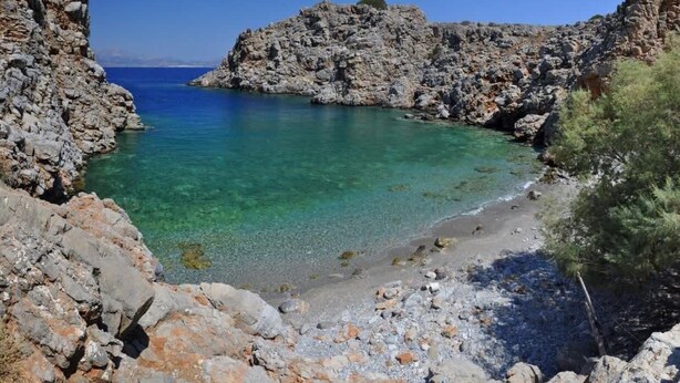 A rocky coastline with clear turquoise water, a pebbly beach, and rugged cliffs.