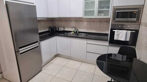 A modern kitchen with white cabinets, a stainless steel refrigerator, a microwave, an oven, and a black dining table with chairs. - Terrace Garden Apartment  (Funchal)