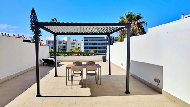 A covered patio area with a table and chairs, a pergola structure, and a view of buildings in the background. - Terrace Garden Apartment  (Funchal)
