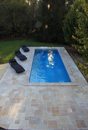 A rectangular swimming pool with a tiled deck, surrounded by greenery and trees.