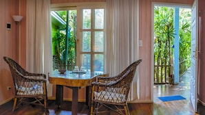 A dining area with a wooden table and wicker chairs, a window with curtains, and a view of a tropical garden through the door. - Secret garden iguazu (Puerto Iguazú)