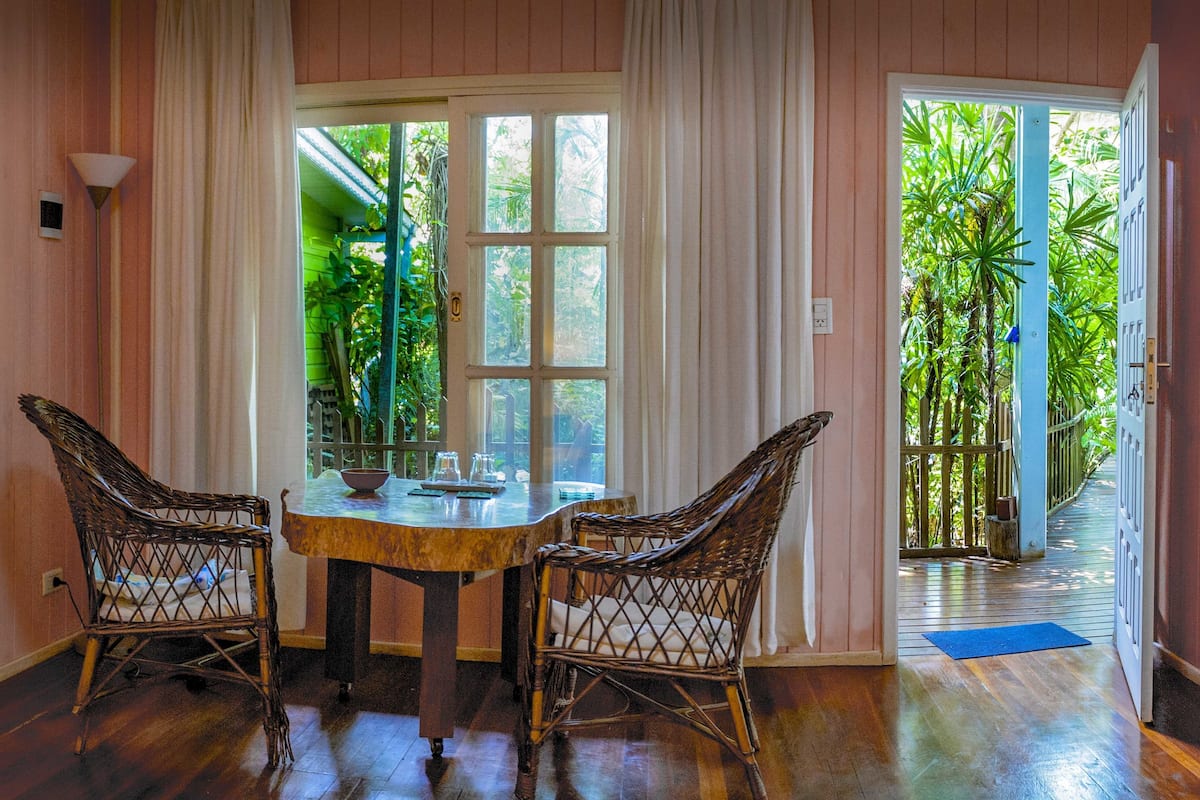 A dining area with a wooden table and wicker chairs, a window with curtains, and a view of a tropical garden through the door.