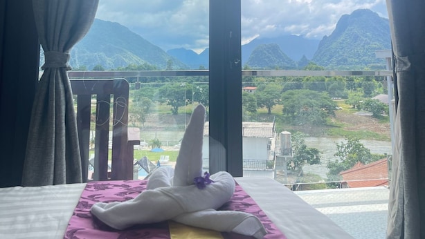A hotel room with a bed, a window with curtains, and a view of mountains and a river. - NuttyPhoy Hotel (Vang Vieng)