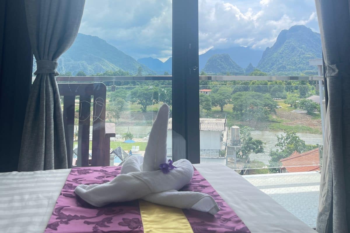 A hotel room with a bed, a window with curtains, and a view of mountains and a river.