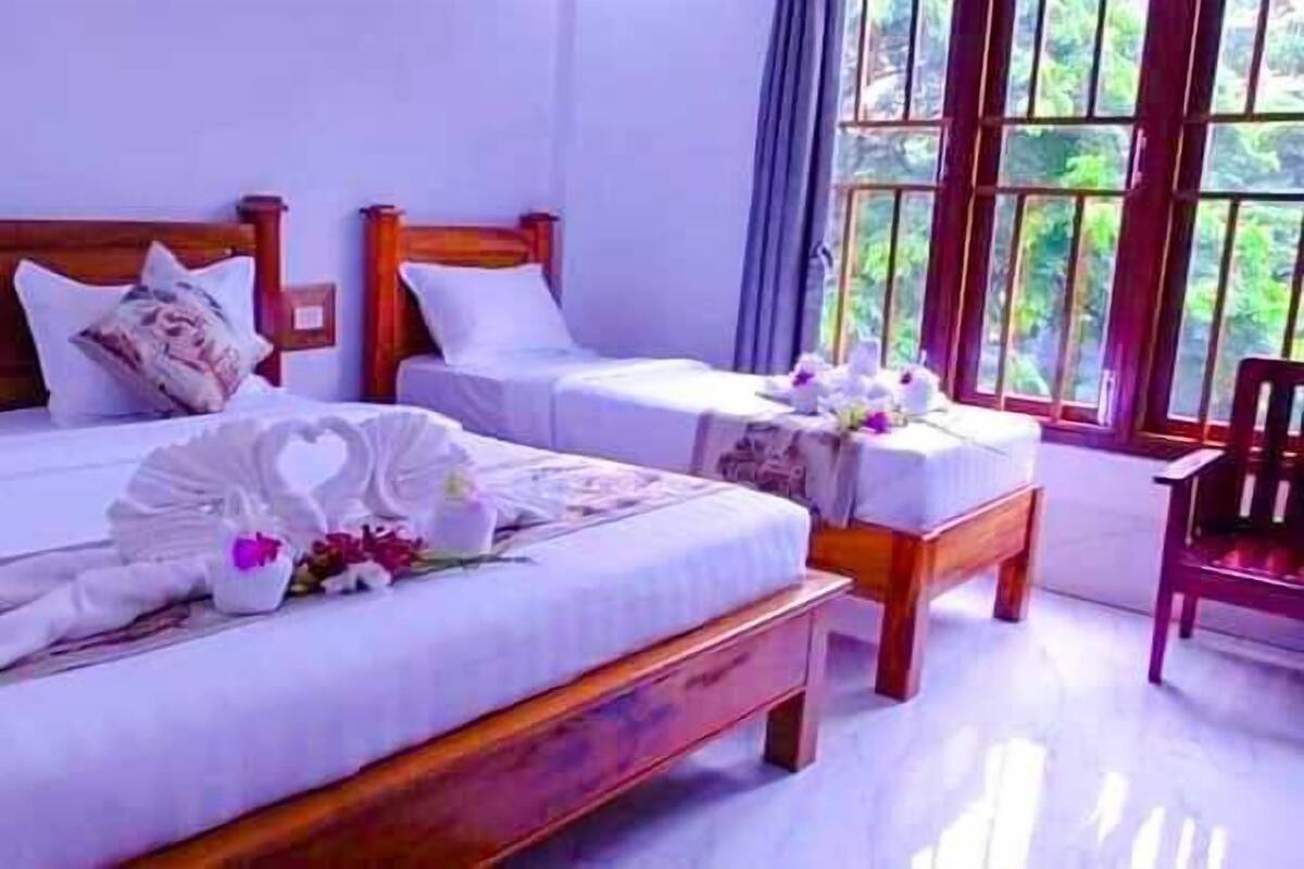 A hotel room with two beds, each with a wooden headboard, white bedding, and decorative pillows. There is a small table with a vase of flowers and a chair.