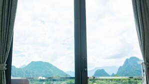 A room with a large window offering a view of green mountains and a cloudy sky. - NuttyPhoy Hotel (Vang Vieng)