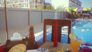 A breakfast setup with eggs, ham, tomatoes, and bread on a table by a poolside with umbrellas and a view of a hotel building. - NuttyPhoy Hotel (Vang Vieng)