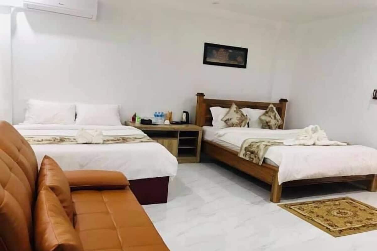A hotel room with two beds, a wooden headboard, a brown leather sofa, a small table with a lamp, and a framed picture on the wall.
