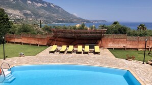 En swimmingpool med liggestole og en træpergola, omgivet af en stenbelagt terrasse og udsigt til hav og bjerge.