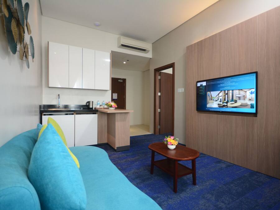 A hotel room with a blue sofa, a small wooden table with a flower vase, a television on the wall, and a kitchenette in the background.