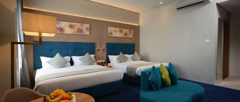 Quarto de hotel com duas camas, área de estar com uma mesa e uma poltrona azul.